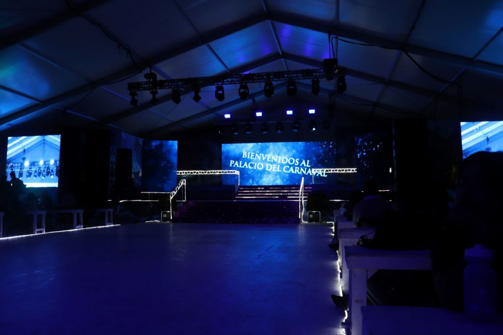 View of the dance performance floor leading to the stage and screen area of Palacio del Carnaval in Cozumel Mexico. The room is lit with purple lighting. The front screens are displaying a Welcome message for arriving guests.
