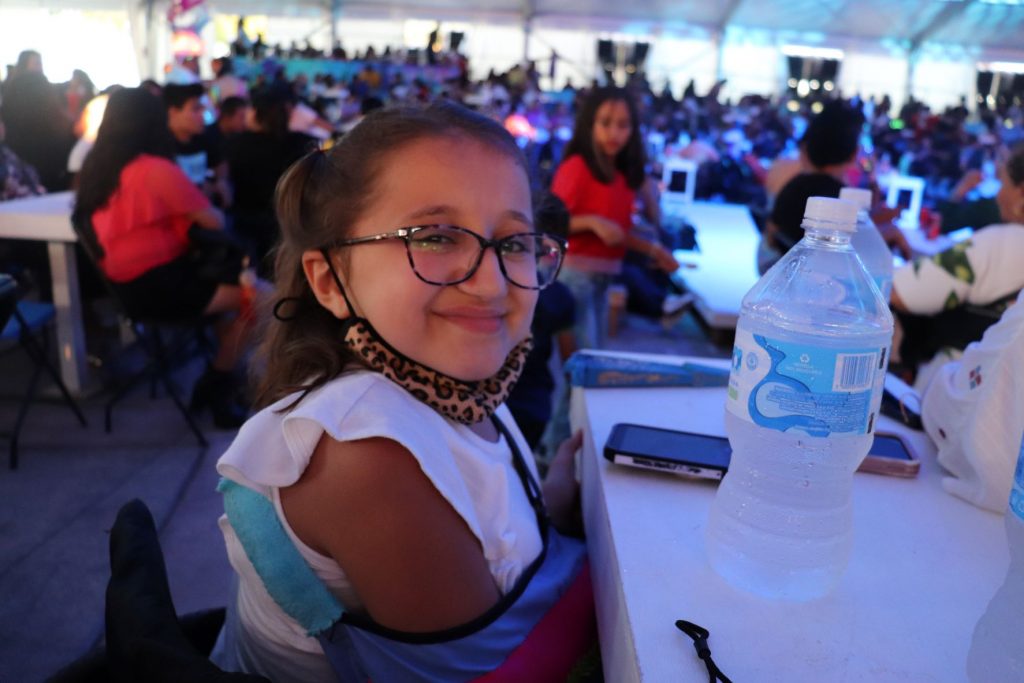 Anna, a teen wheelchair user with long brown hair and wearing glasses, is smiling at the camera as she waits for a performance to begin. She has a bottle of water set on the table next to her alongside her cellphone. She is wearing a white ruffled t-shirt and has an arm in a blue medical sling, as she waits for a Carnaval dance performance in Cozumel Mexico.