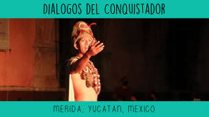 A latino male actor in his 30's is dressed in traditional Mayan costume while speaking on an outdoor stage. His his and raised as he passionately tells a story.