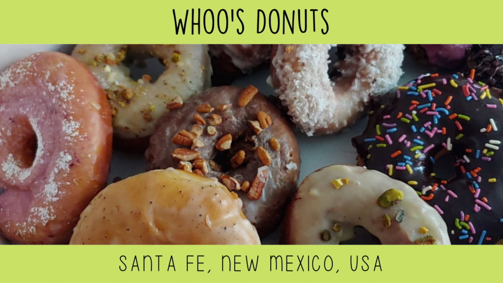 A close up photo of a variety of doughnuts. A lemon glaze with poppyseeds, a chocolate frosted with multicolor sprinkles, a white iced donut with chopped pistachios. a glazed cake donut with chopped pecans, a vanilla frosted donut dipped in coconut and a glazed donut with red cake peeking through and a light dusting of powdered sugar.