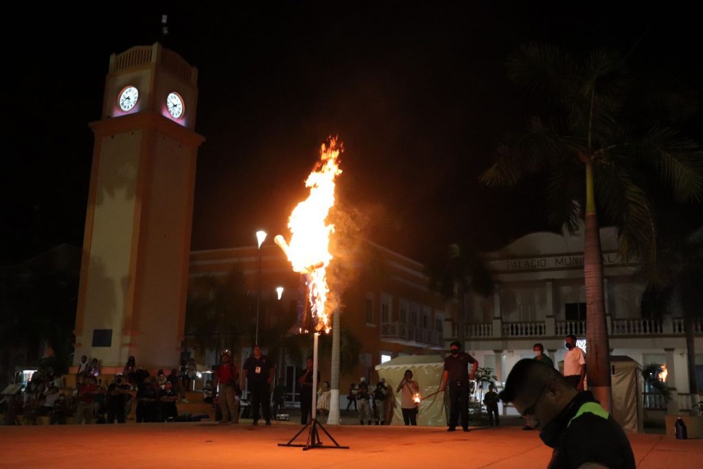The Cozumel town buildings and clock tower surround the stage where officials are standing by as the Juan Carnaval effigy on display burns. There are some spectators visible, but most are kept at a distance for fire safety.