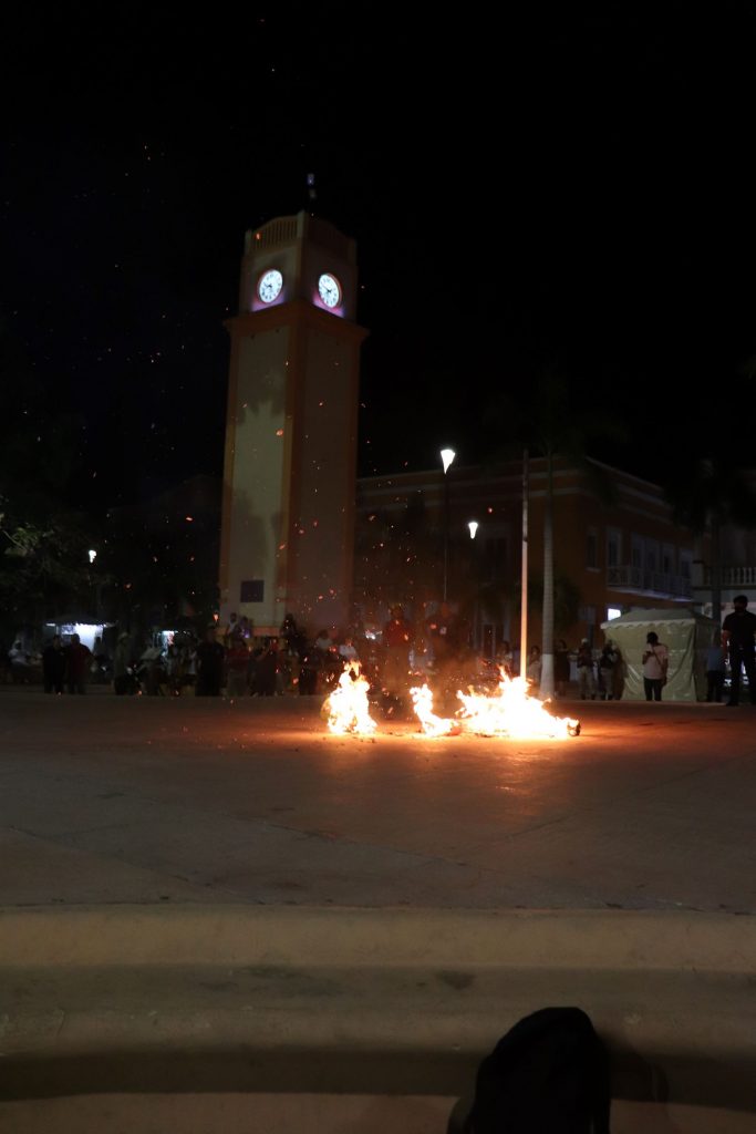 Pieces of the Juan Carnaval effigy are in flames on the stage at Parque Benito Juarez in Cozumel Mexico. Spectators are visible in the dim light and the iconic clock tower stands in the background.